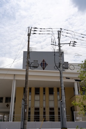 Two utility poles with multiple electrical wires are positioned in front of a modern building with a diamond-shaped window. The building has a yellow and white facade, and the sky is lightly clouded.
