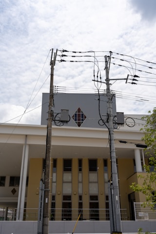 Two utility poles with multiple electrical wires are positioned in front of a modern building with a diamond-shaped window. The building has a yellow and white facade, and the sky is lightly clouded.