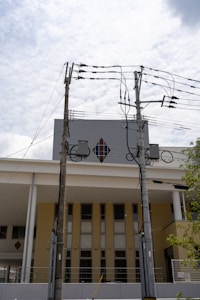 Two utility poles with multiple electrical wires are positioned in front of a modern building with a diamond-shaped window. The building has a yellow and white facade, and the sky is lightly clouded.