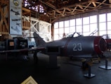A vintage aircraft is displayed inside a dimly lit hangar with wooden beams. The aircraft, marked with the number 23 and a red star, is metallic with red accents. Large windows line the wall in the background, allowing some light to filter in. Posters and other aircraft-related displays are visible around the hangar.