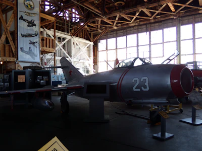 A vintage aircraft is displayed inside a dimly lit hangar with wooden beams. The aircraft, marked with the number 23 and a red star, is metallic with red accents. Large windows line the wall in the background, allowing some light to filter in. Posters and other aircraft-related displays are visible around the hangar.