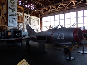 A vintage aircraft is displayed inside a dimly lit hangar with wooden beams. The aircraft, marked with the number 23 and a red star, is metallic with red accents. Large windows line the wall in the background, allowing some light to filter in. Posters and other aircraft-related displays are visible around the hangar.