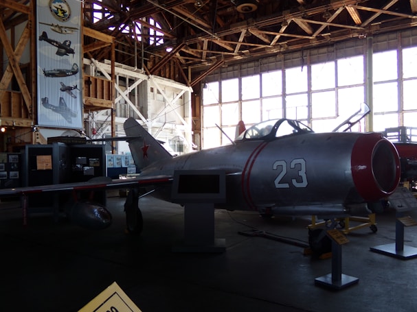 A vintage aircraft is displayed inside a dimly lit hangar with wooden beams. The aircraft, marked with the number 23 and a red star, is metallic with red accents. Large windows line the wall in the background, allowing some light to filter in. Posters and other aircraft-related displays are visible around the hangar.