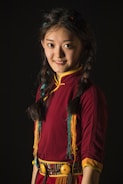 A young woman with braided hair, wearing a traditional dress with colorful fringes and intricate patterns, is smiling gently. The deep red and yellow accents in her attire contrast with the dark background.