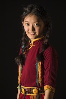 A young woman with braided hair, wearing a traditional dress with colorful fringes and intricate patterns, is smiling gently. The deep red and yellow accents in her attire contrast with the dark background.
