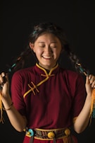 A smiling person with long braided hair wearing a traditional maroon and yellow outfit, holding their braids with both hands against a dark background.