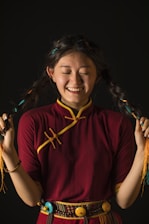 A smiling person with long braided hair wearing a traditional maroon and yellow outfit, holding their braids with both hands against a dark background.