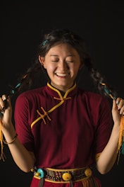 A smiling person with long braided hair wearing a traditional maroon and yellow outfit, holding their braids with both hands against a dark background.