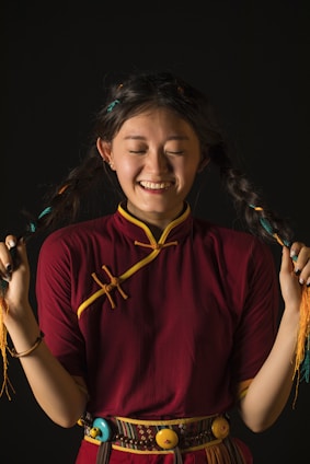 A smiling person with long braided hair wearing a traditional maroon and yellow outfit, holding their braids with both hands against a dark background.