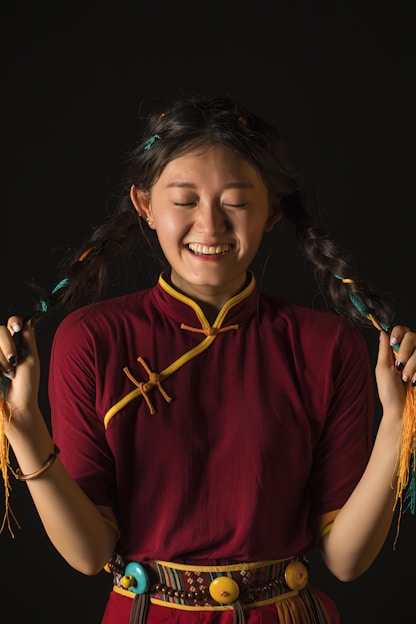 A smiling person with long braided hair wearing a traditional maroon and yellow outfit, holding their braids with both hands against a dark background.