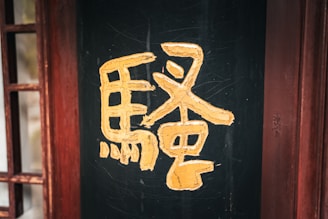 A large yellow Chinese character is painted on a dark, possibly black, wooden surface. The wood has a textured appearance, and there are distressed reddish wooden frames on the side, providing a rustic and traditional ambiance.