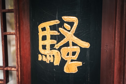A large yellow Chinese character is painted on a dark, possibly black, wooden surface. The wood has a textured appearance, and there are distressed reddish wooden frames on the side, providing a rustic and traditional ambiance.