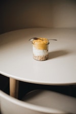 A small jar containing a layered dessert is placed on a round white table. The dessert appears to consist of granola, cream or yogurt, and a yellow fruit puree. A small spoon is resting on top of the jar.