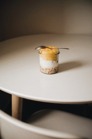 A small jar containing a layered dessert is placed on a round white table. The dessert appears to consist of granola, cream or yogurt, and a yellow fruit puree. A small spoon is resting on top of the jar.