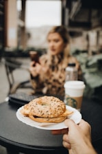 A hand holds a white plate with a sesame seed bagel sandwich on it, in an outdoor café setting. In the blurred background, a person with long hair is seated at a table, handling a smartphone and sitting next to a cup with a sleeve, likely from a popular coffee shop.