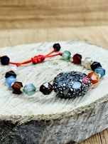 A bracelet made of multifaceted beads in various colors is placed on a piece of wooden surface. The beads are connected by a red string, with a textured, darker bead as the central piece. The background is a blurred wooden texture.