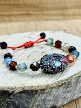 A bracelet made of multifaceted beads in various colors is placed on a piece of wooden surface. The beads are connected by a red string, with a textured, darker bead as the central piece. The background is a blurred wooden texture.