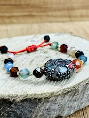 A bracelet made of multifaceted beads in various colors is placed on a piece of wooden surface. The beads are connected by a red string, with a textured, darker bead as the central piece. The background is a blurred wooden texture.