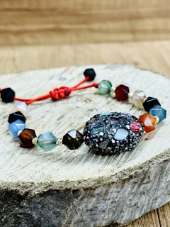 A bracelet made of multifaceted beads in various colors is placed on a piece of wooden surface. The beads are connected by a red string, with a textured, darker bead as the central piece. The background is a blurred wooden texture.