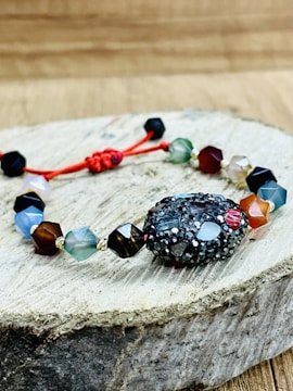 A bracelet made of multifaceted beads in various colors is placed on a piece of wooden surface. The beads are connected by a red string, with a textured, darker bead as the central piece. The background is a blurred wooden texture.