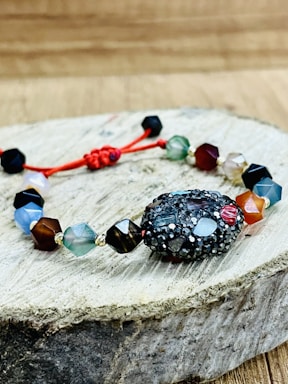 A bracelet made of multifaceted beads in various colors is placed on a piece of wooden surface. The beads are connected by a red string, with a textured, darker bead as the central piece. The background is a blurred wooden texture.