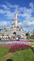A fairy-tale castle with pink walls, blue spires, and golden accents stands against a bright blue sky. In the foreground, vibrant flowers in shades of pink and purple are planted in a garden. People are walking towards the entrance of the castle, enjoying the sunny day.