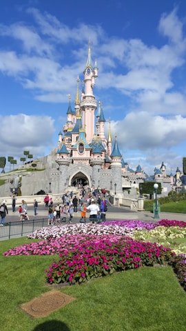 A fairy-tale castle with pink walls, blue spires, and golden accents stands against a bright blue sky. In the foreground, vibrant flowers in shades of pink and purple are planted in a garden. People are walking towards the entrance of the castle, enjoying the sunny day.