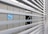 Slatted blinds partially obscure the view of an industrial structure with metal casing. A blue circular sign with 'BAC' is visible. The environment appears to be an industrial setting with elements of metal and machinery.