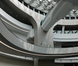 A modern architectural interior featuring curved concrete structures and sleek lines. Light filters through large windows onto the minimalist design, creating patterns and shadows. The space is open and airy with multiple levels visible.