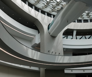 A modern architectural interior featuring curved concrete structures and sleek lines. Light filters through large windows onto the minimalist design, creating patterns and shadows. The space is open and airy with multiple levels visible.