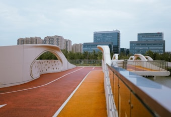 A modern pedestrian bridge with an artistic design featuring white arching structures and perforated patterns. The bridge is set against a background of contemporary office buildings and residential apartments, surrounded by greenery and a clear sky.