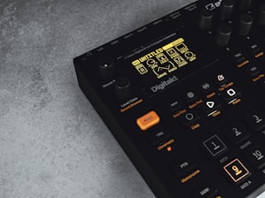 A digital music production device with a display screen showing menu options and buttons. The interface features various controls like knobs and illuminated buttons, labeled with functions including 'Trig,' 'Tap Tempo,' and 'Copy.' The device has a sleek black design with an industrial vibe, complemented by a textured grey surface underneath.