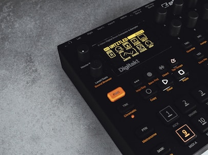 A digital music production device with a display screen showing menu options and buttons. The interface features various controls like knobs and illuminated buttons, labeled with functions including 'Trig,' 'Tap Tempo,' and 'Copy.' The device has a sleek black design with an industrial vibe, complemented by a textured grey surface underneath.