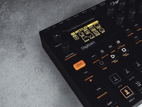 A digital music production device with a display screen showing menu options and buttons. The interface features various controls like knobs and illuminated buttons, labeled with functions including 'Trig,' 'Tap Tempo,' and 'Copy.' The device has a sleek black design with an industrial vibe, complemented by a textured grey surface underneath.