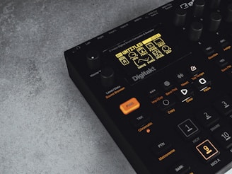A digital music production device with a display screen showing menu options and buttons. The interface features various controls like knobs and illuminated buttons, labeled with functions including 'Trig,' 'Tap Tempo,' and 'Copy.' The device has a sleek black design with an industrial vibe, complemented by a textured grey surface underneath.