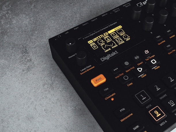 A digital music production device with a display screen showing menu options and buttons. The interface features various controls like knobs and illuminated buttons, labeled with functions including 'Trig,' 'Tap Tempo,' and 'Copy.' The device has a sleek black design with an industrial vibe, complemented by a textured grey surface underneath.