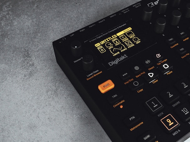 A digital music production device with a display screen showing menu options and buttons. The interface features various controls like knobs and illuminated buttons, labeled with functions including 'Trig,' 'Tap Tempo,' and 'Copy.' The device has a sleek black design with an industrial vibe, complemented by a textured grey surface underneath.