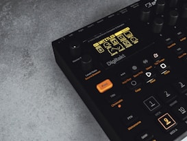 A digital music production device with a display screen showing menu options and buttons. The interface features various controls like knobs and illuminated buttons, labeled with functions including 'Trig,' 'Tap Tempo,' and 'Copy.' The device has a sleek black design with an industrial vibe, complemented by a textured grey surface underneath.