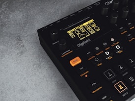 A digital music production device with a display screen showing menu options and buttons. The interface features various controls like knobs and illuminated buttons, labeled with functions including 'Trig,' 'Tap Tempo,' and 'Copy.' The device has a sleek black design with an industrial vibe, complemented by a textured grey surface underneath.