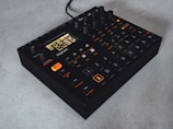 A compact, black electronic music sampler with multiple dials and buttons on its surface. The front panel features an illuminated LCD screen with a menu and various settings, with several buttons labeled for different functions. The device is sitting on a light-colored surface.