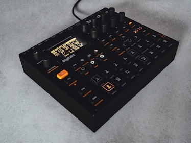 A compact, black electronic music sampler with multiple dials and buttons on its surface. The front panel features an illuminated LCD screen with a menu and various settings, with several buttons labeled for different functions. The device is sitting on a light-colored surface.
