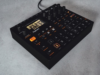A compact, black electronic music sampler with multiple dials and buttons on its surface. The front panel features an illuminated LCD screen with a menu and various settings, with several buttons labeled for different functions. The device is sitting on a light-colored surface.