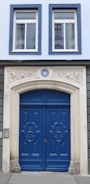 A building facade featuring a pair of elegant blue wooden doors adorned with intricate carvings. Above the doors is a decorative stone arch with the number 16 at the center. The facade includes two large windows with white frames, and the building's exterior is painted in a combination of light blue and grey tones.