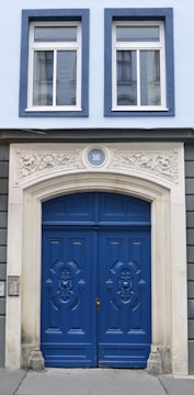 A building facade featuring a pair of elegant blue wooden doors adorned with intricate carvings. Above the doors is a decorative stone arch with the number 16 at the center. The facade includes two large windows with white frames, and the building's exterior is painted in a combination of light blue and grey tones.