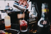 A hand is pouring a red liquid from a cocktail shaker into a glass filled with crushed ice. Next to the glass, there is a bottle of Johnnie Walker Black Label whiskey, partially filled. An orange squeeze bottle is also visible on the side.