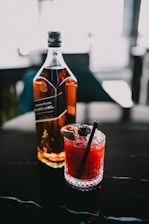 A bottle of whiskey stands next to a glass filled with a vibrant red cocktail, garnished with a blackberry and a black straw. The scene is set on a dark, shiny surface with blurred background elements.