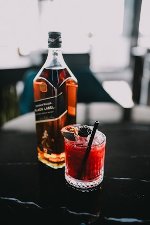A bottle of whiskey stands next to a glass filled with a vibrant red cocktail, garnished with a blackberry and a black straw. The scene is set on a dark, shiny surface with blurred background elements.