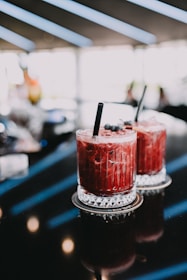 Two red cocktails with black straws sit on coasters on a glossy, dark surface. The drinks have a textured surface and are garnished with small berries on top. The background is softly blurred, indicating a modern, ambient setting with streaks of light.