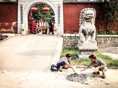 Two individuals kneel near a pile of ashes on the ground, seemingly engaged in some form of ritual or offering, next to a large stone lion statue. In the background, a group of people dressed in ceremonial attire stand under a stone archway adorned with traditional red lanterns. The setting appears to be a cultural or religious site with a reddish-brown wall and lush greenery nearby.