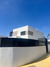 A modern, minimalist house with clean lines and geometric shapes. The exterior features a combination of white walls and light wooden panels. A high black fence surrounds the property, and there is a clear blue sky in the background.